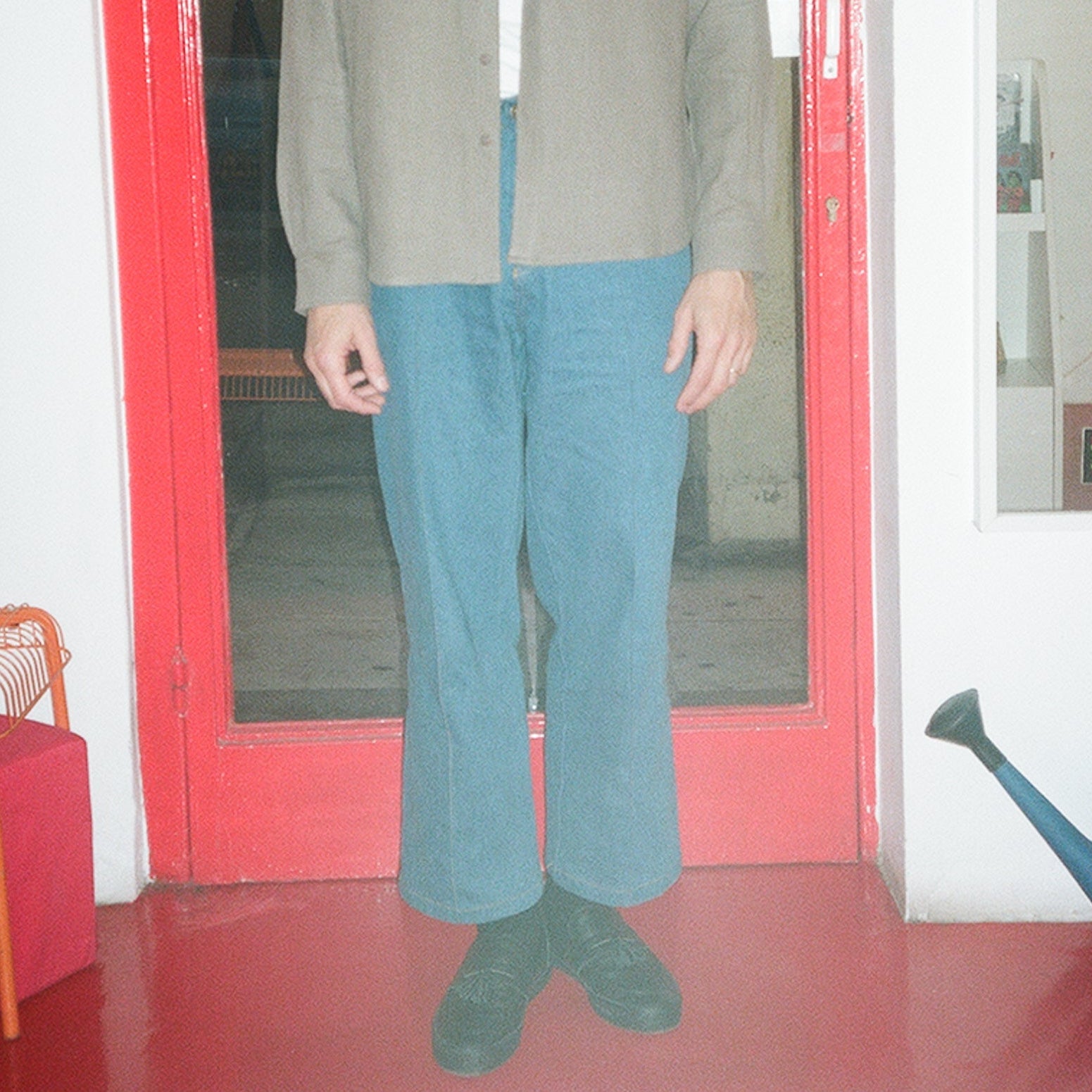 Person wearing a beige jacket and blue jeans standing in front of a red door.